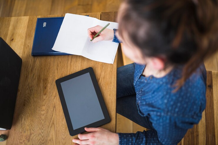 Woman writing notes on paper with digital tablet over wooden desk